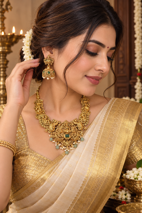 Elegant matte gold-finish Temple Jewellery Set featuring an intricate Goddess Lakshmi motif with beautifully detailed elephant designs and green stone accents. Comes with matching jhumka earrings(slight variation in color & design may occur)