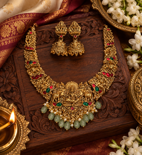 Elegant matte gold-finish Temple Jewellery Set featuring an intricate Goddess Lakshmi motif with beautifully detailed elephant designs and green stone accents. Comes with matching jhumka earrings(slight variation in color & design may occur)
