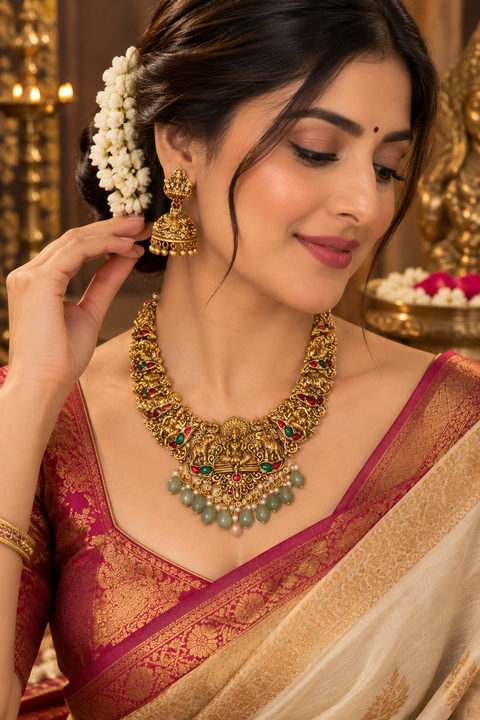 Elegant matte gold-finish Temple Jewellery Set featuring an intricate Goddess Lakshmi motif with beautifully detailed elephant designs and green stone accents. Comes with matching jhumka earrings(slight variation in color & design may occur)
