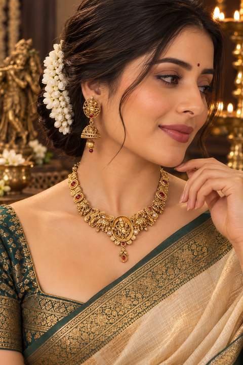Elegant matte gold-finish Temple Jewellery Set featuring an intricate Radha Krishna motif with beautifully detailed elephant designs and Ruby stone accents. Comes with matching jhumka earrings(slight variation in color & design may occur)